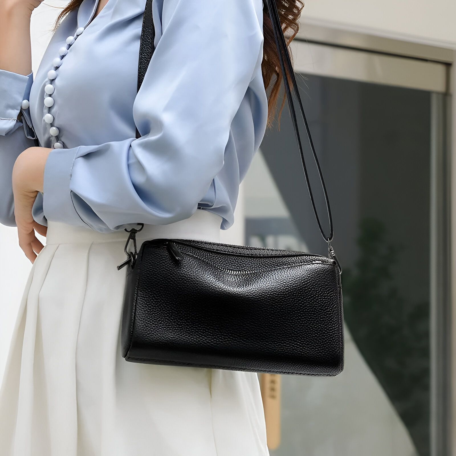 Une femme portant une chemise bleue et une jupe blanche porte un sac à bandoulière noir en cuir.