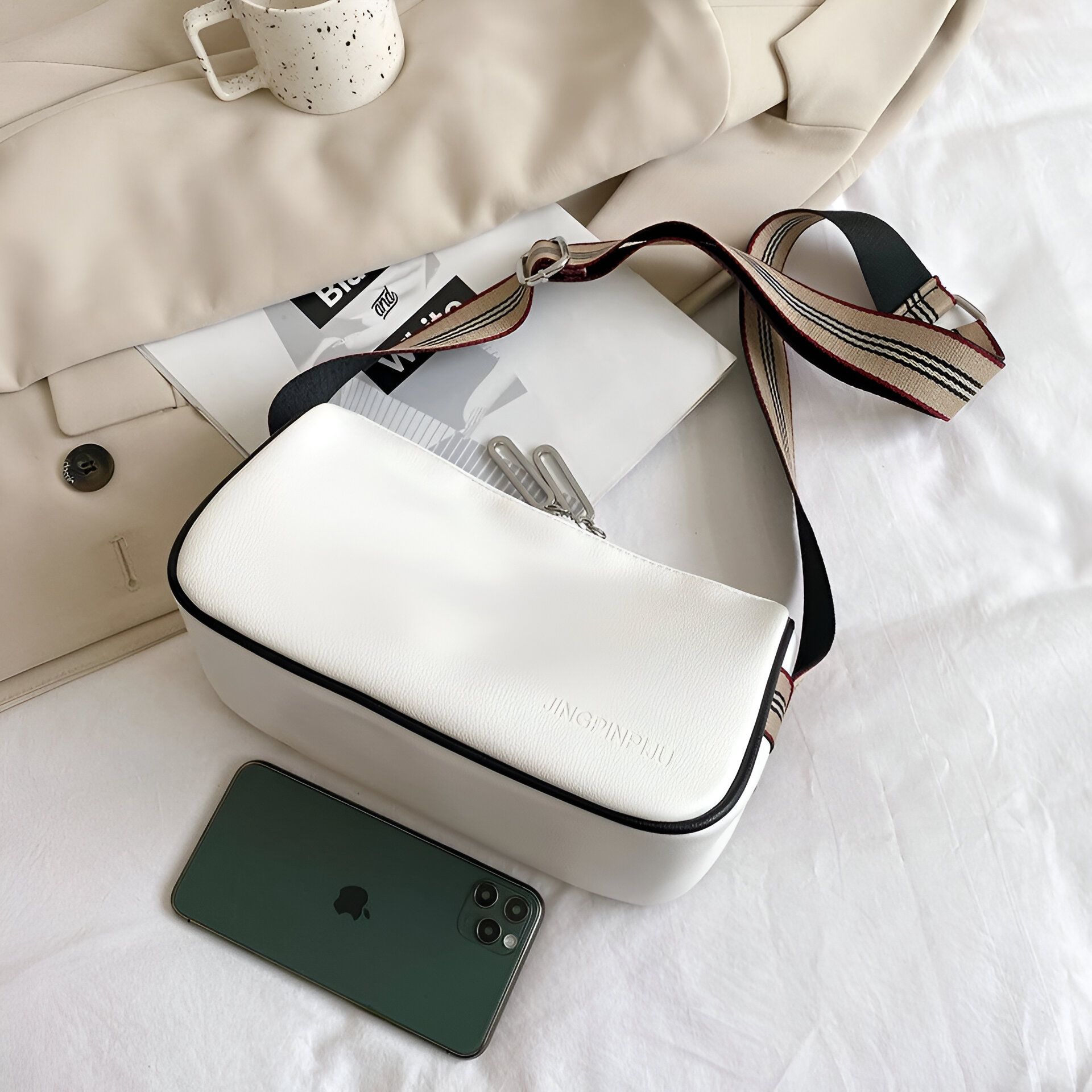 Un sac à main blanc avec une bandoulière noire et blanche, posé sur un lit blanc. Un téléphone portable vert est à côté du sac.