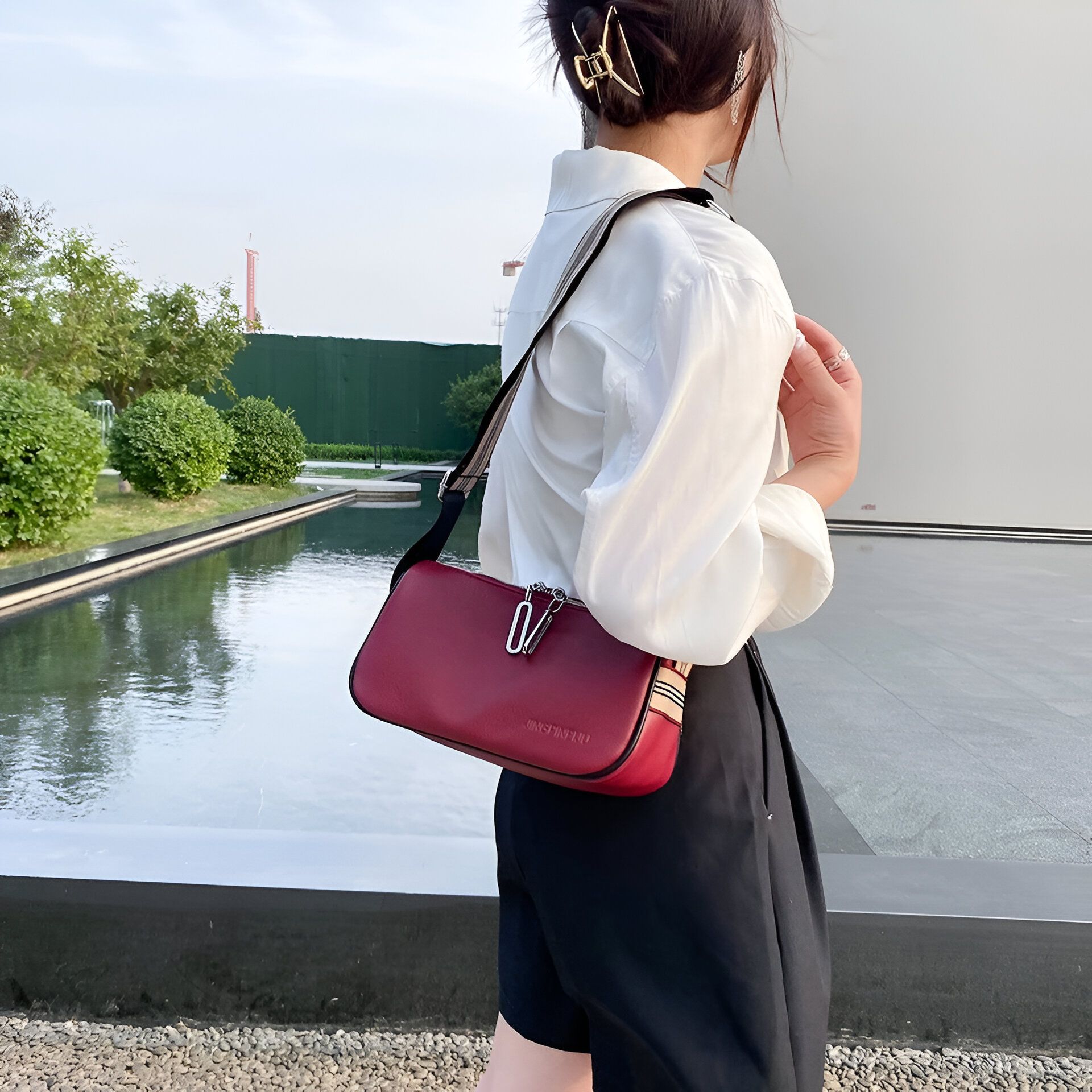 Une femme portant un chemisier blanc et un short noir, avec un sac à main rouge à bandoulière. Elle est debout devant un bassin d'eau.