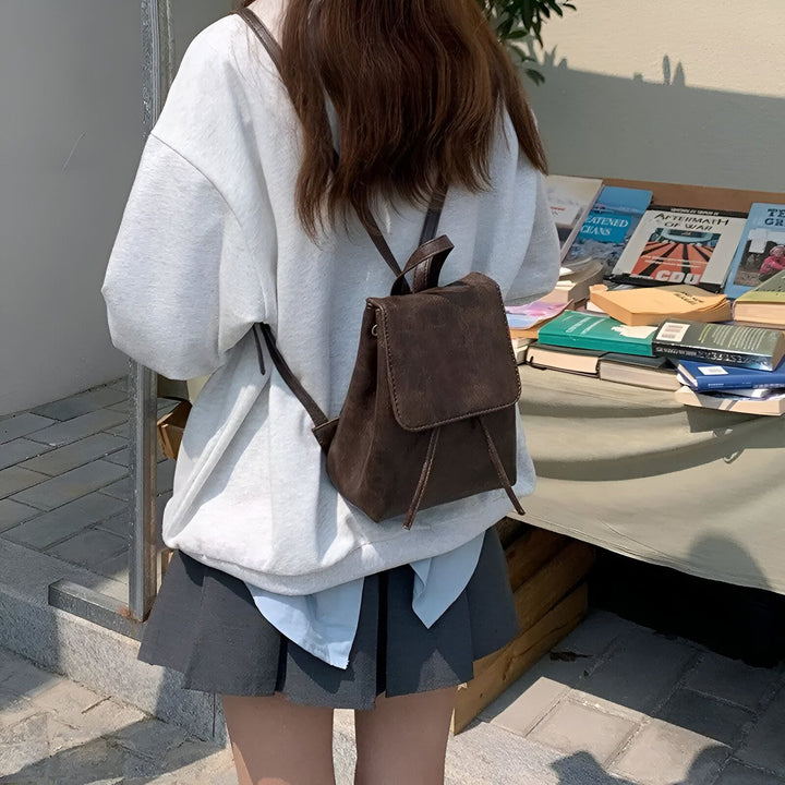 Personne de dos avec sweat gris, jupe et mini sac à dos près d'une table de livres., 46194001805536,46194001838304