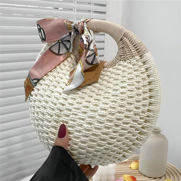 Sac rond en paille tressée avec poignée et foulard rose et blanc, tenu à la main., 46193945706720,46193945739488,46193945772256,46193945805024,46193945837792