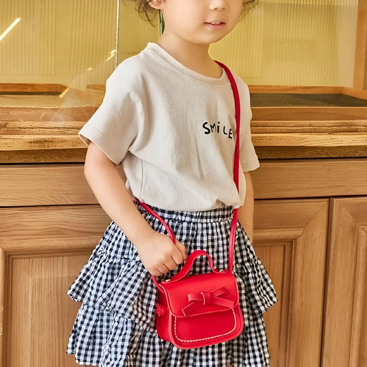 Fille tenant petit sac rouge avec nœud, jupe à carreaux et t-shirt., 46193988698336,46193988731104,46193988796640,46193988829408,46193988862176,46193988894944,46193988927712