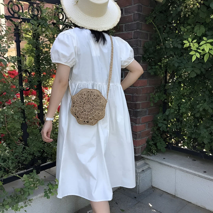 Femme de dos en robe blanche, chapeau de paille et sac rond en paille, à côté d'un mur de briques et de roses., 46193946034400,46193946067168