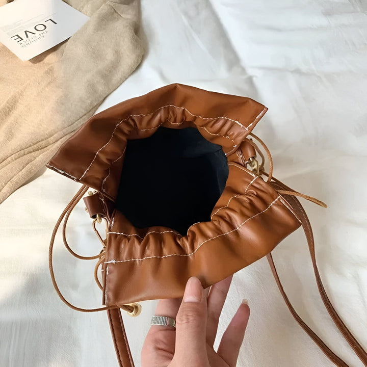 Sac seau en cuir marron avec cordon de serrage, intérieur noir et coutures blanches, tenu ouvert par une main., 46194011308256,46194011373792