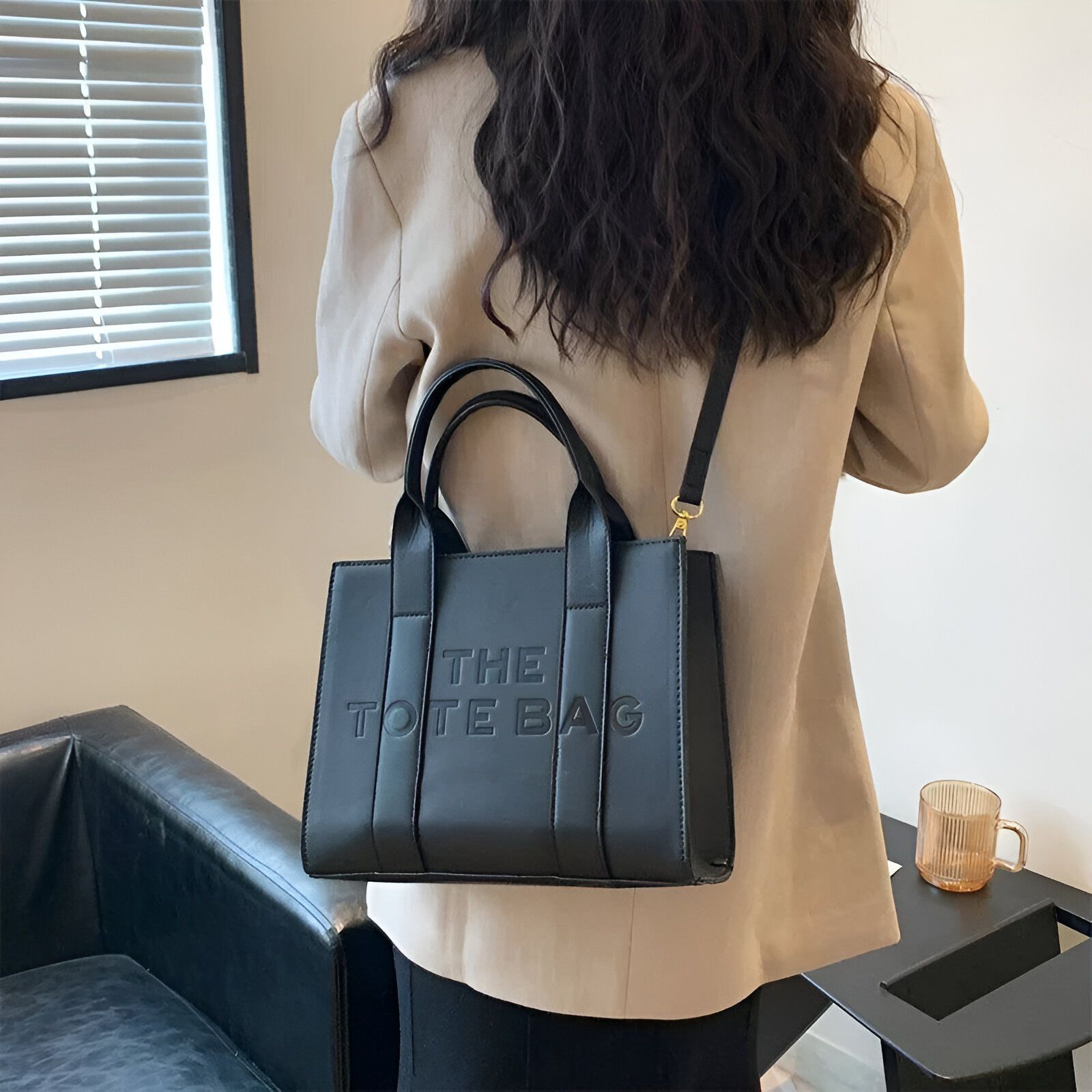 Une femme portant un sac à main noir en cuir avec l'inscription "The Tote Bag" en lettres dorées.