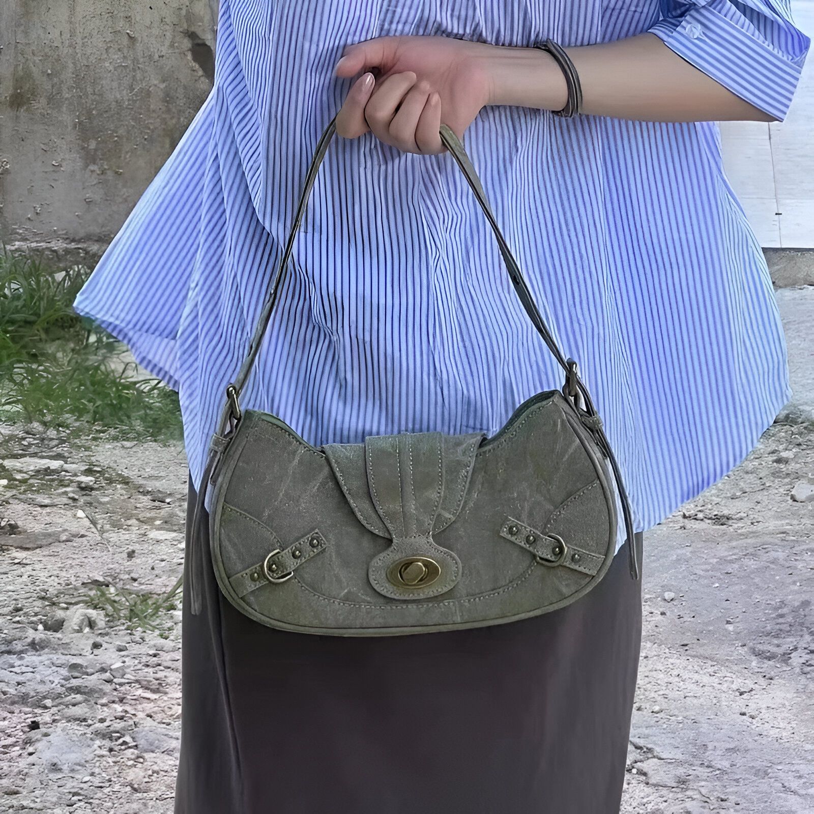 Une femme porte un sac à main en toile de couleur vert foncé avec une fermeture dorée. Le sac est porté sur l'épaule et la femme porte une chemise bleue à rayures.