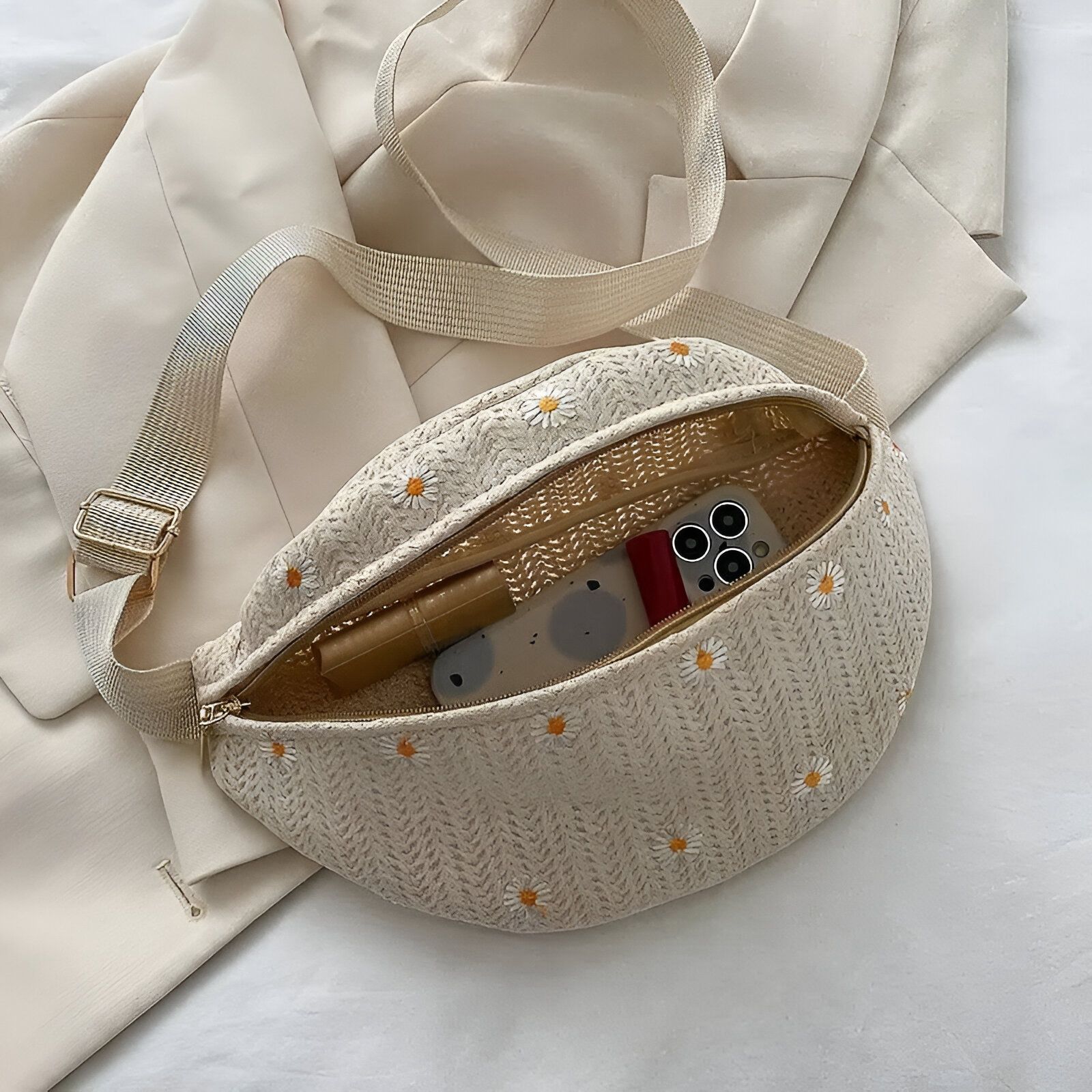 Sac banane en tissu beige avec des fleurs blanches brodées. Il est ouvert et on peut voir l'intérieur du sac avec un téléphone portable, un étui à lunettes et un rouge à lèvres.