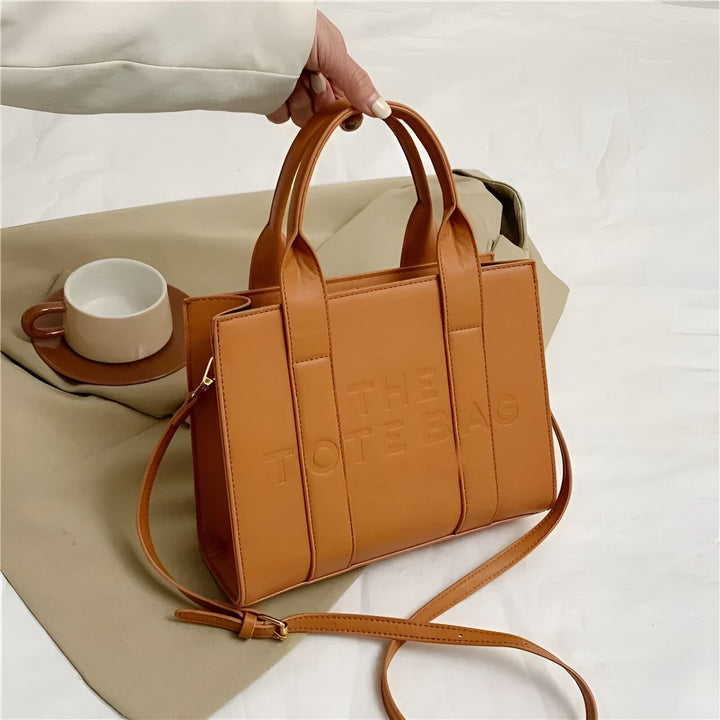 Une main tient un sac tote bag marron gravé "THE TOTE BAG"., 46193877811424,46193877844192,46193877876960,46193877909728,46193877942496,46193877975264,46193878008032,46193878040800