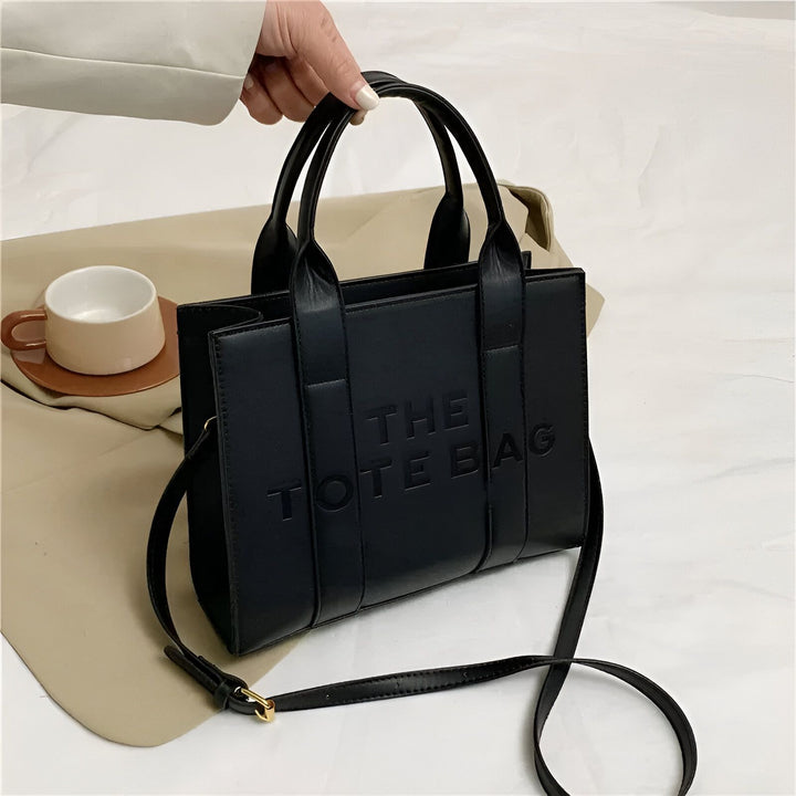 Main tenant un sac tote bag noir "THE TOTE BAG" avec bandoulière., 46193877811424,46193877844192,46193877876960,46193877909728,46193877942496,46193877975264,46193878008032,46193878040800