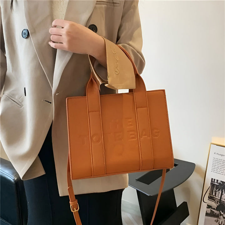 Sac cabas orange en cuir avec inscription "THE TOTE BAG" porté par une personne en blazer., 46193877811424,46193877844192,46193877876960,46193877909728,46193877942496,46193877975264,46193878008032,46193878040800