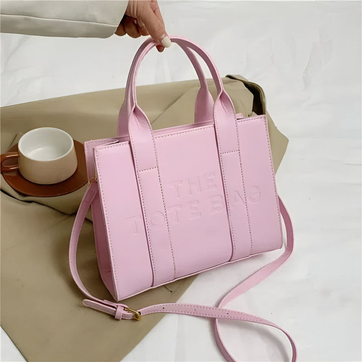 Main tenant un sac tote bag rose pâle avec "THE TOTE BAG" embossé et bandoulière., 46193877811424,46193877844192,46193877876960,46193877909728,46193877942496,46193877975264,46193878008032,46193878040800