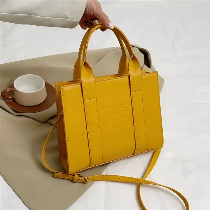 Sac cabas jaune "THE TOTE BAG" tenu par une main, avec bandoulière., 46193877811424,46193877844192,46193877876960,46193877909728,46193877942496,46193877975264,46193878008032,46193878040800