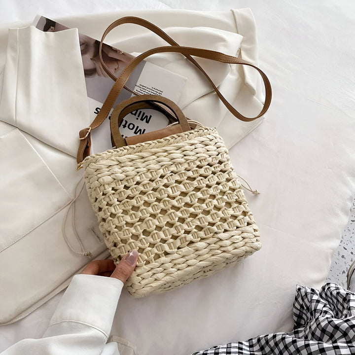 Sac en paille beige avec lanière en cuir marron et poignées en bois, tenu par une main, sur fond clair., 46193946722528,46193946755296