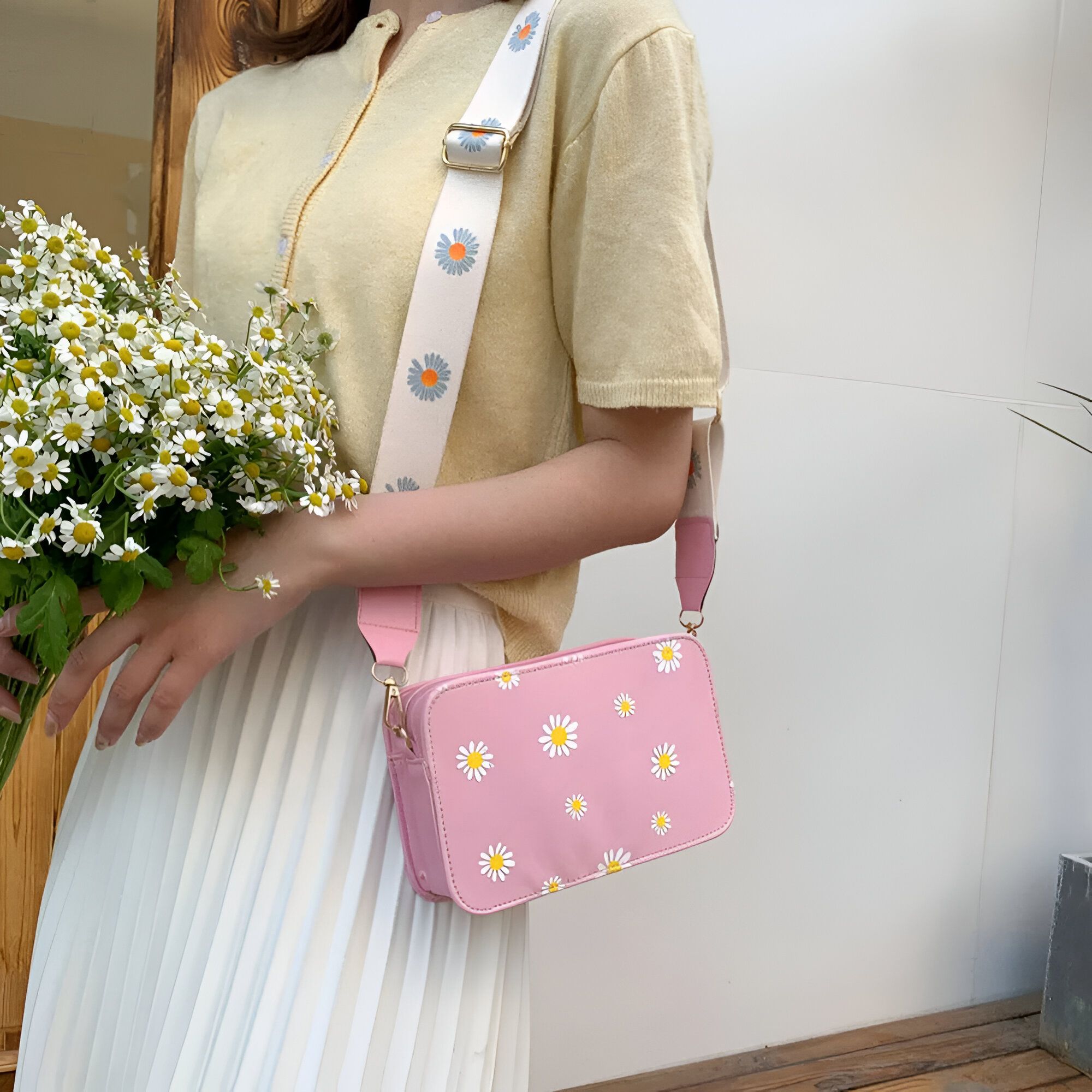 Une femme porte un sac à main rose avec un motif de marguerites. Le sac à main a une bandoulière blanche avec un motif de marguerites. La femme porte un haut jaune et une jupe blanche.