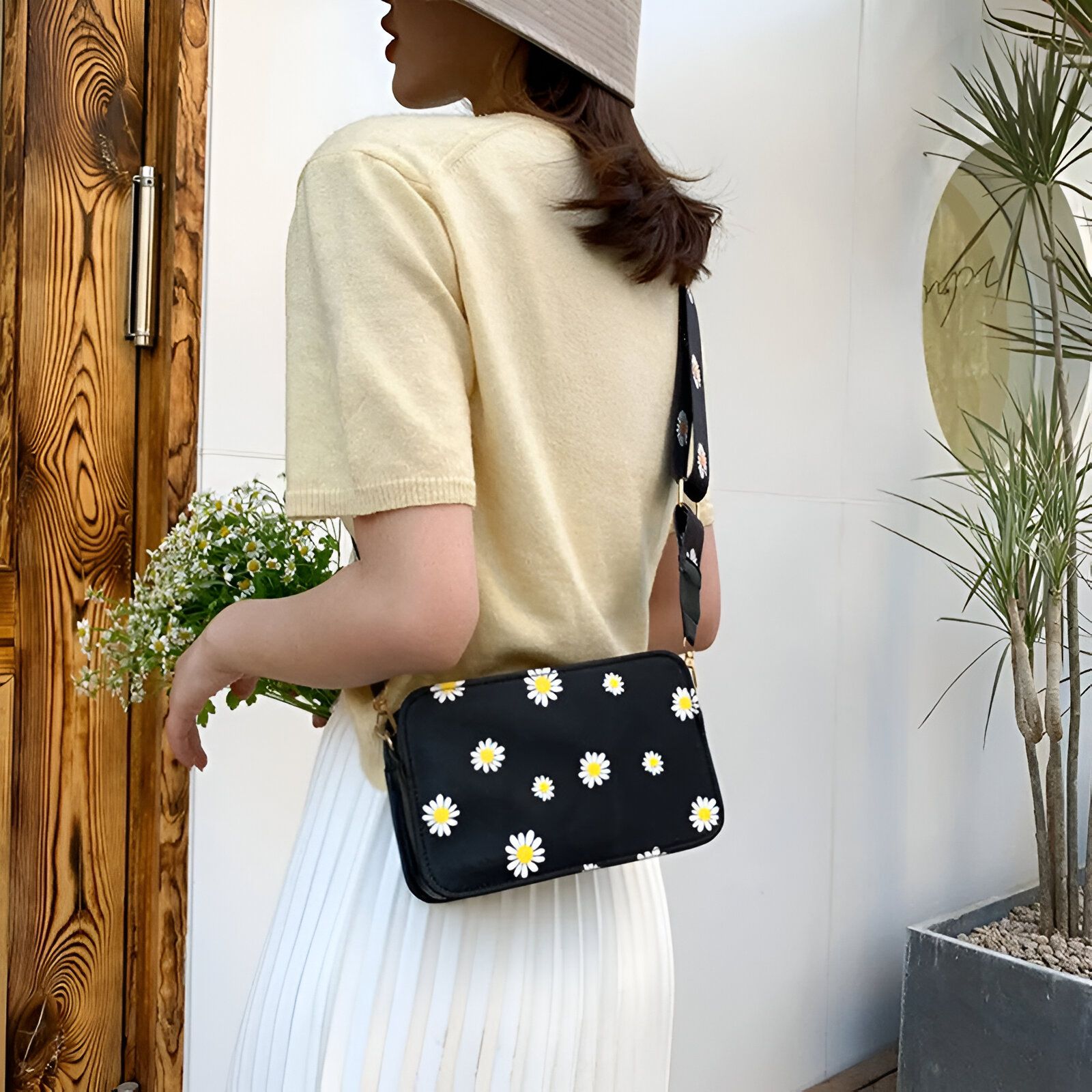 Une femme porte un sac à main noir avec un motif de marguerites jaunes.  Le sac est porté en bandoulière.  Elle porte un t-shirt jaune et une jupe blanche.  Elle porte également un chapeau.