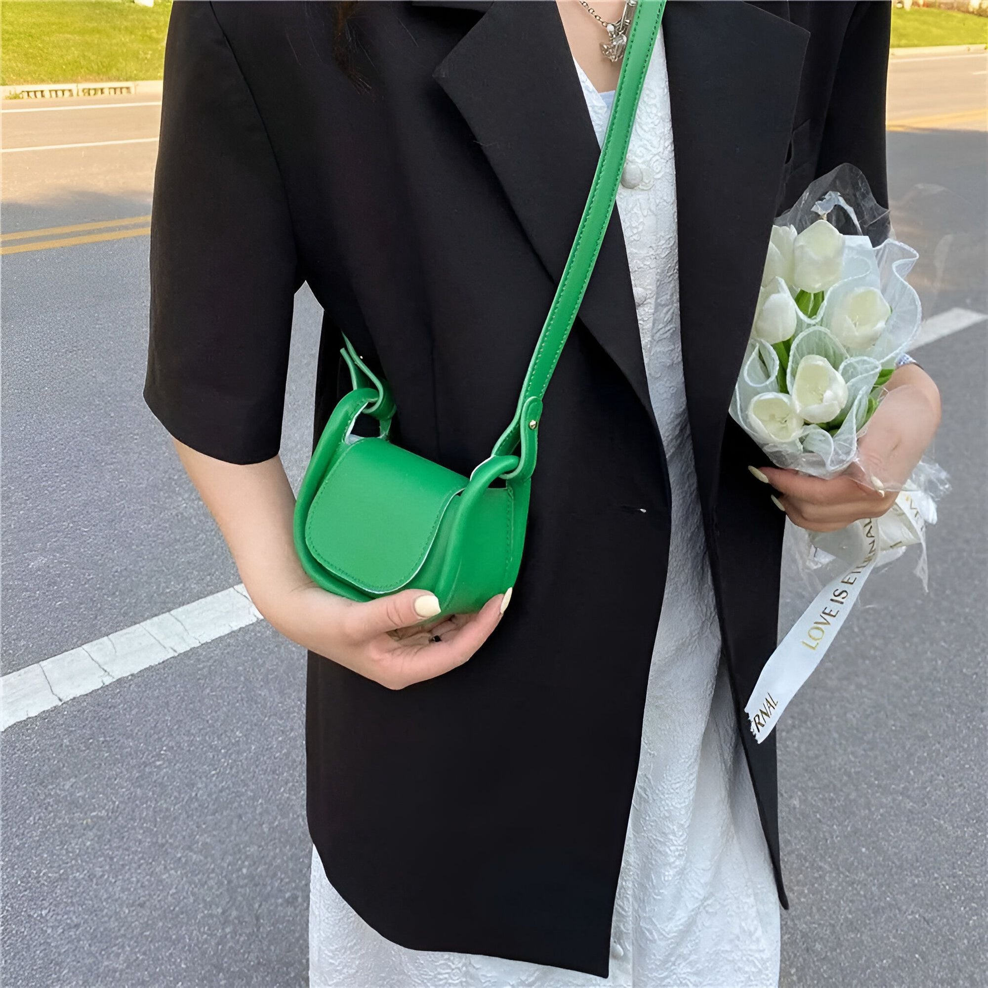 Une femme portant un blazer noir et un petit sac à main vert.  Elle tient également un bouquet de fleurs blanches.