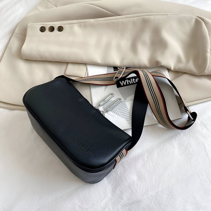 Sac à bandoulière noir avec sangle rayée posé sur un blazer beige., 46266441433312,46266441466080,46266441498848