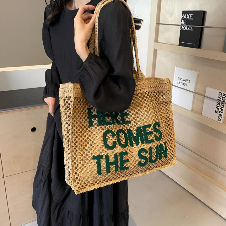 Sac fourre-tout en paille ajourée avec l'inscription verte "HERE COMES THE SUN" porté par une personne., 46193944723680,46193944756448
