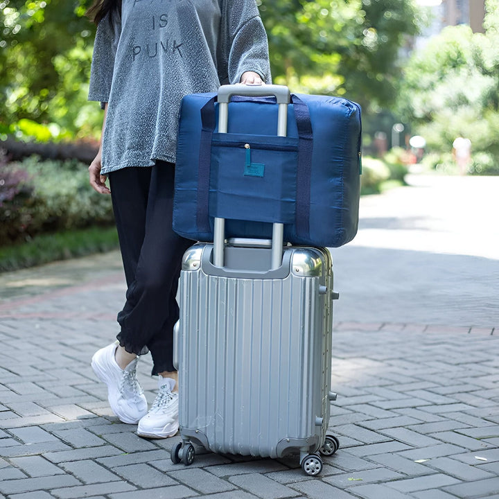 Sac de voyage pliable bleu sur une valise roulante argentée tenue par une personne., 46193919852768,46193919885536,46193919918304,46193919951072,46193919983840