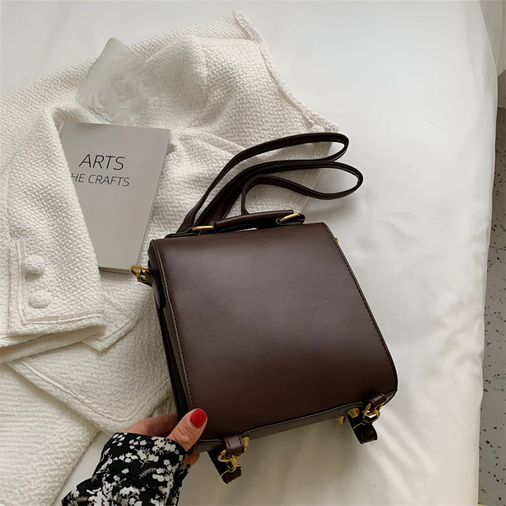 Sac à bandoulière marron avec détails dorés sur fond blanc texturé, près d'un livre., 46194003312864,46194003345632,46194003378400,46194003411168