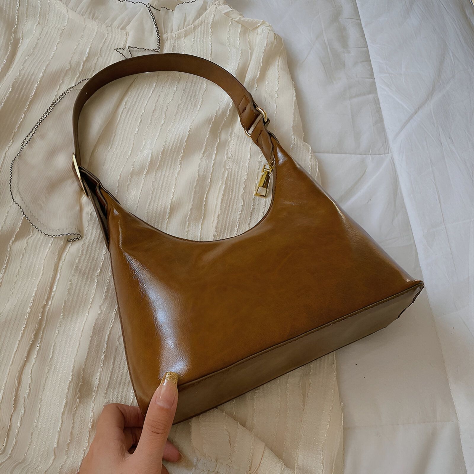 Un sac à main marron en cuir avec une bandoulière en cuir.  Le sac est posé sur un lit blanc.