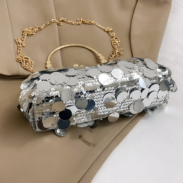 Sac pochette argenté à sequins brillants, avec chaîne et anse dorées, posé sur un tissu beige., 46194028478688,46194028511456