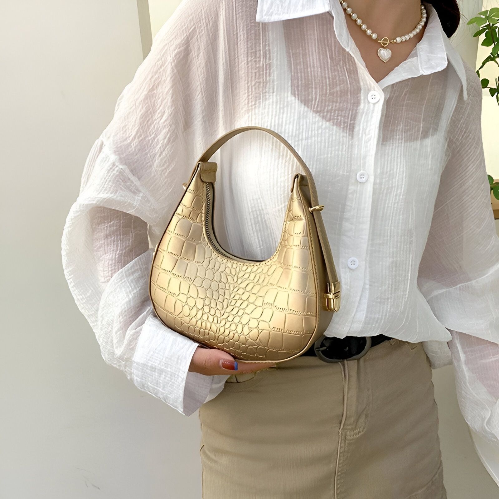 Un sac à main en forme de croissant de lune doré avec une texture de crocodile, tenu par une femme portant une chemise blanche et un short beige.