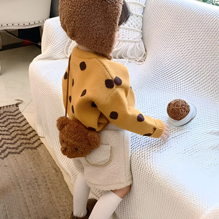 Enfant de dos avec pull jaune à pois, short doux, chapeau marron et sac nounours., 46193981587680,46193981620448