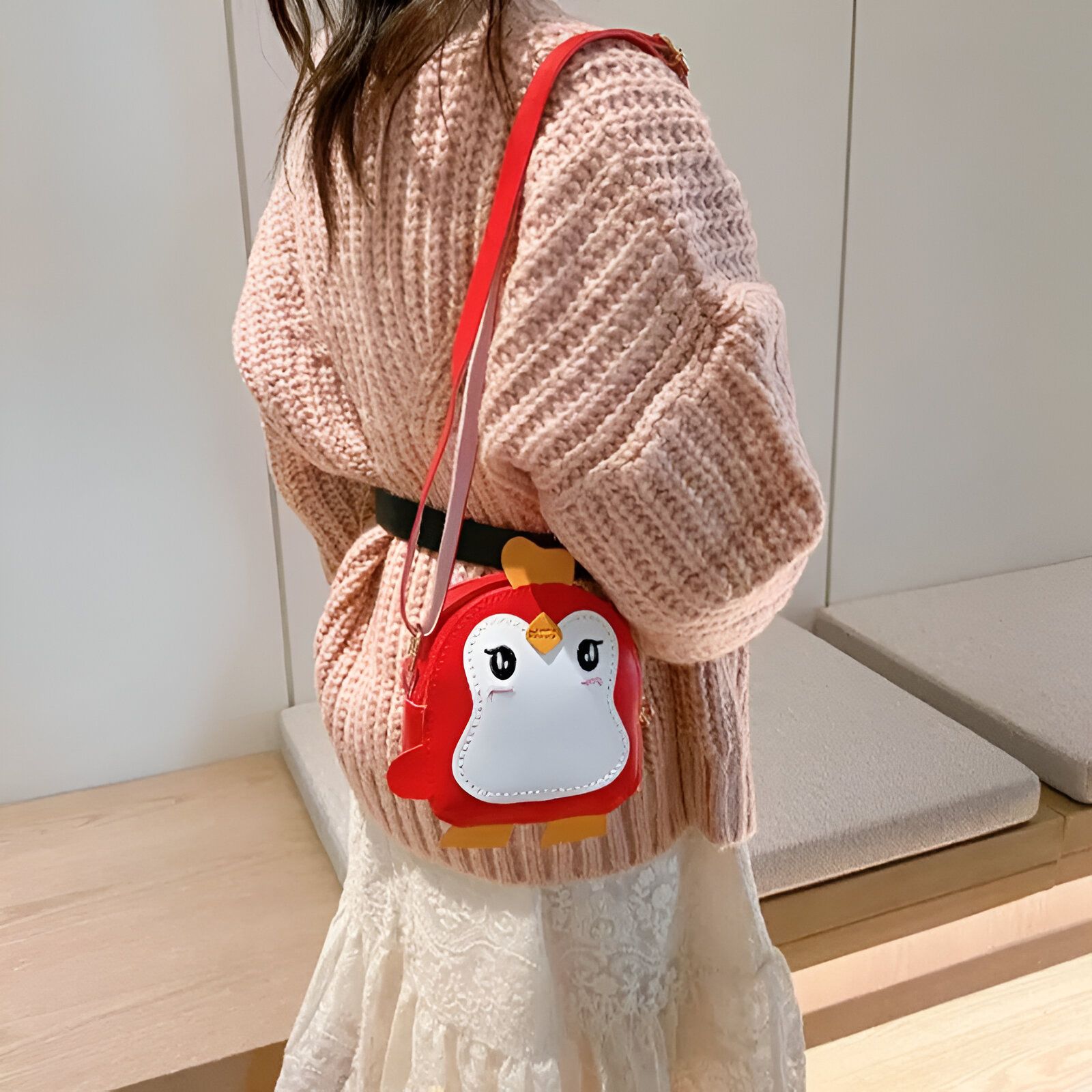 Une jeune fille portant un chandail rose tricoté et une jupe blanche porte un sac à main en forme de pingouin rouge.