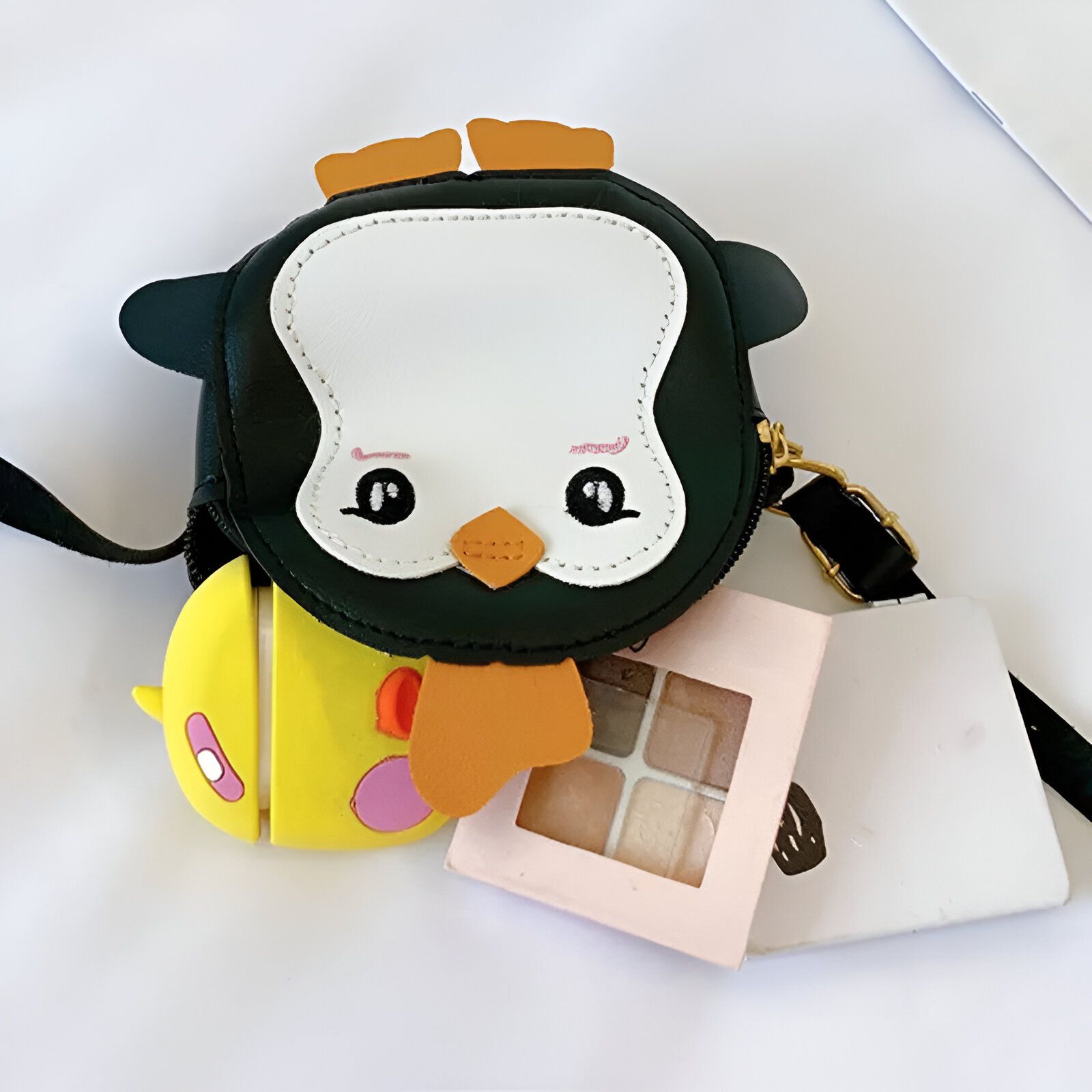 Un petit sac à main en forme de pingouin noir avec un bec orange, des yeux noirs et un visage blanc. Le sac est sur une surface blanche avec un petit étui jaune et une palette de maquillage rose à côté de lui.  Le sac est doté d'une bandoulière noire.