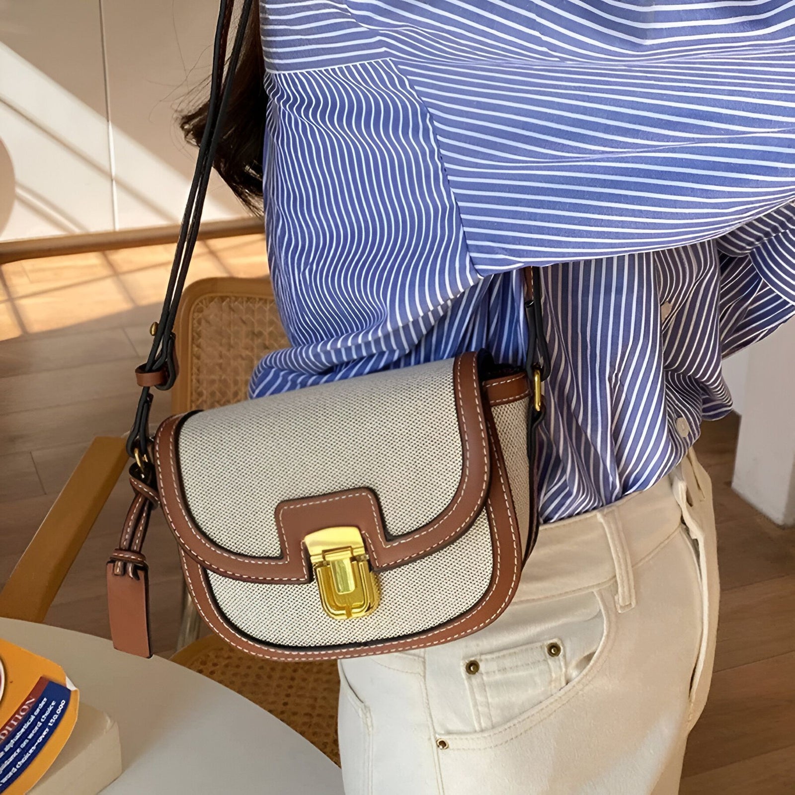 Un gros plan d'une femme portant un sac à main en toile beige avec des accents bruns.  Le sac à main a une fermeture à boucle dorée.  La femme porte une chemise bleue à rayures et un pantalon blanc.