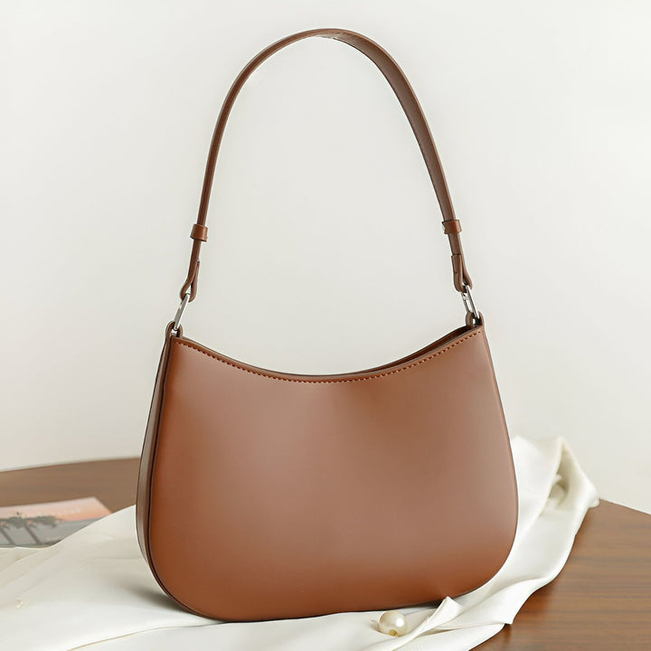 Sac baguette en cuir marron avec anse, posé sur une table en bois avec tissu blanc et perle., 46193850056928
