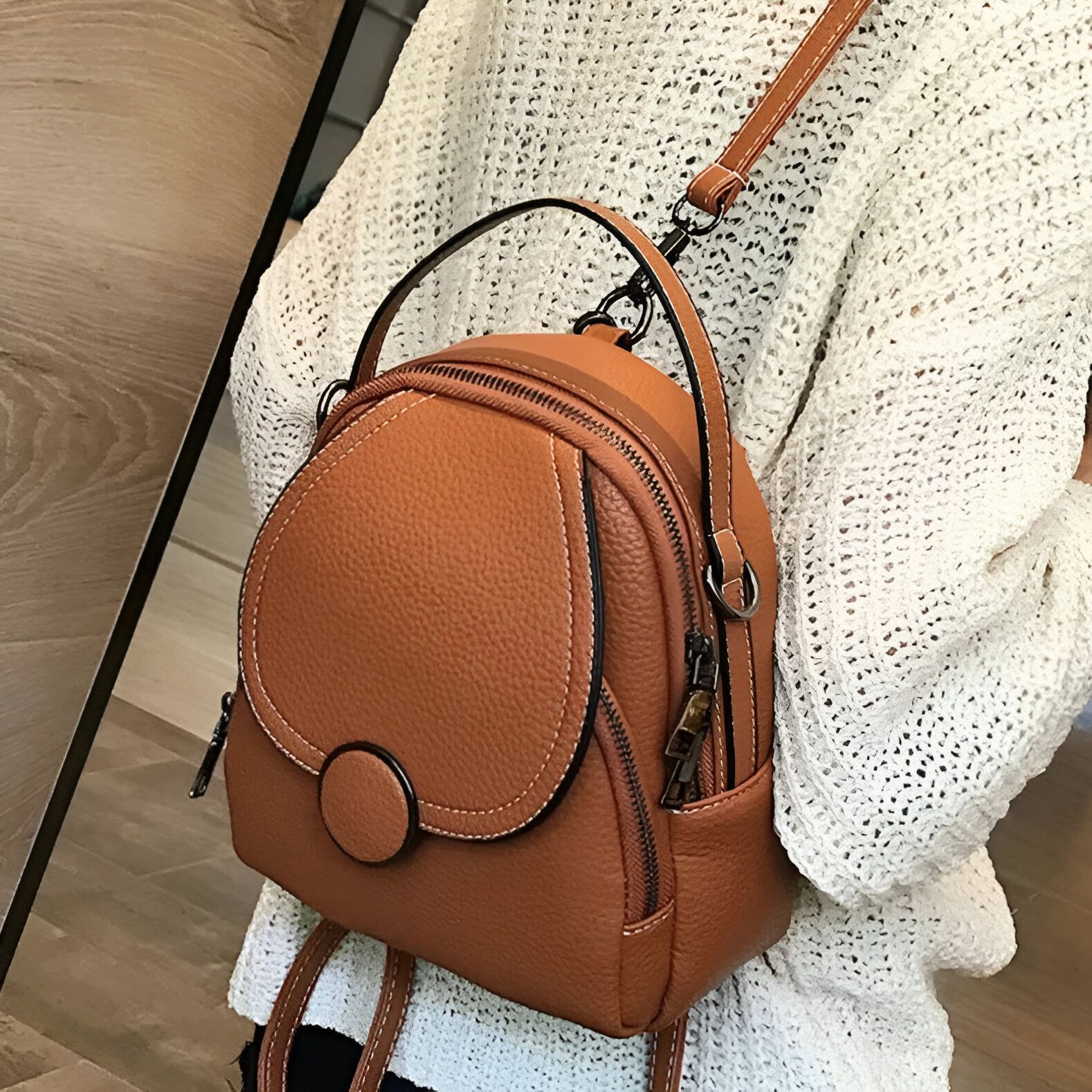 Un sac à dos en cuir brun porté par une femme. Le sac à dos a une fermeture éclair sur le dessus et une sangle sur chaque épaule.