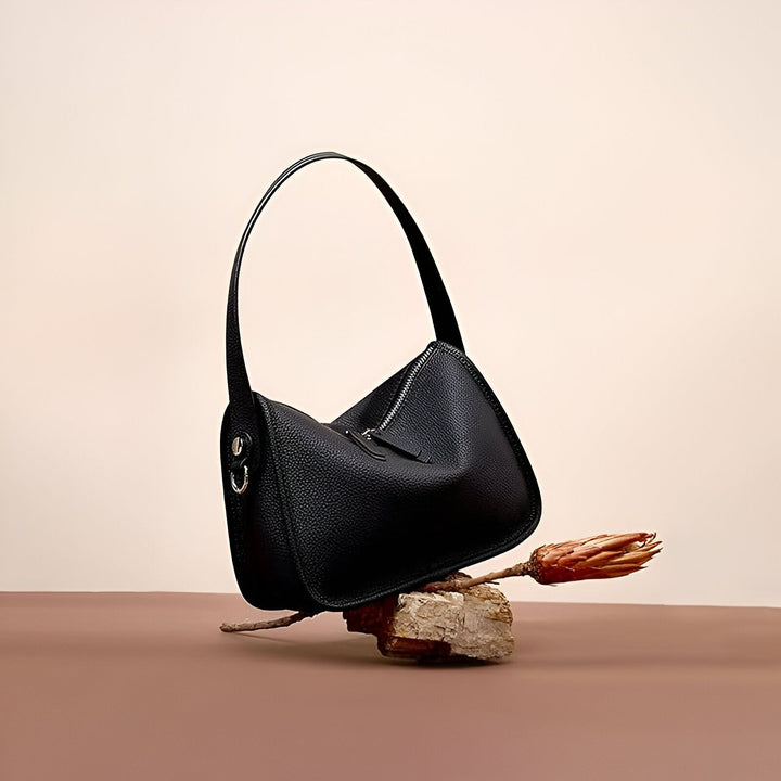 Sac en cuir noir à l'épaule posé sur une roche avec fleur séchée., 46193861820640