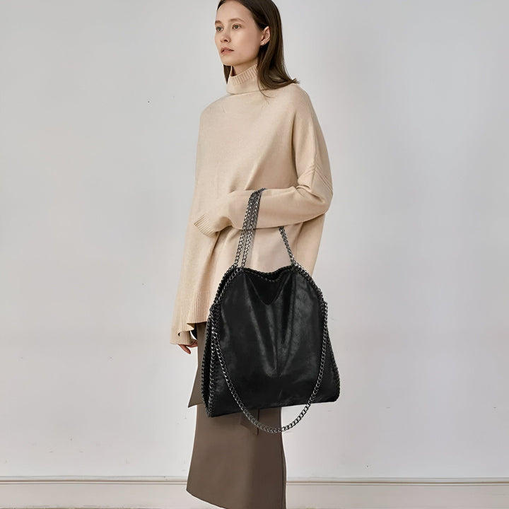 Femme tenant un grand sac fourre-tout noir avec chaînes argentées, pull beige et jupe marron., 46193949245664,46193949278432,46193949311200