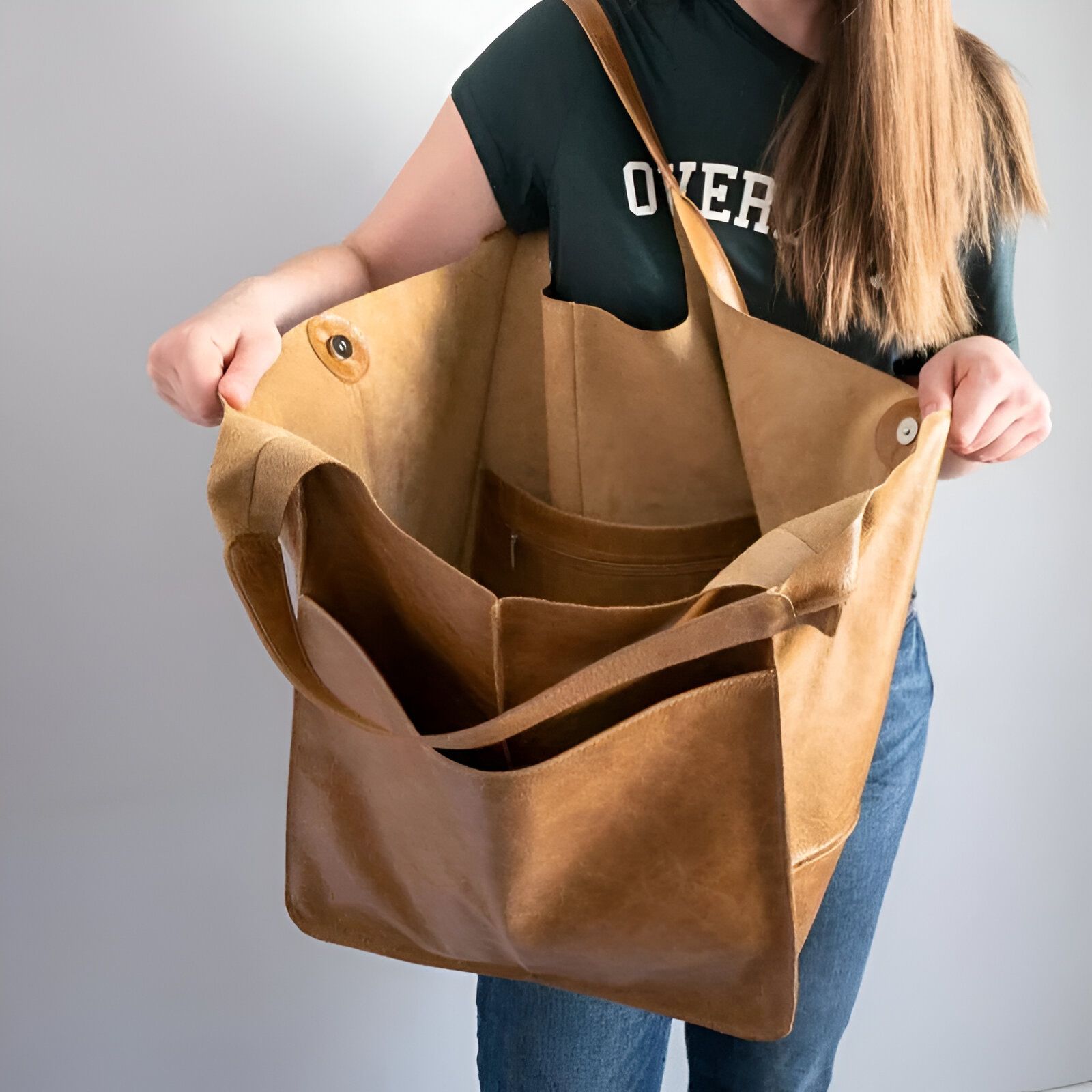 Un grand sac à main en cuir brun ouvert pour montrer son intérieur.  Il y a des poches intérieures et une doublure en tissu.  Le sac est porté par une femme.  Le sac est parfait pour le shopping, le travail ou les voyages.