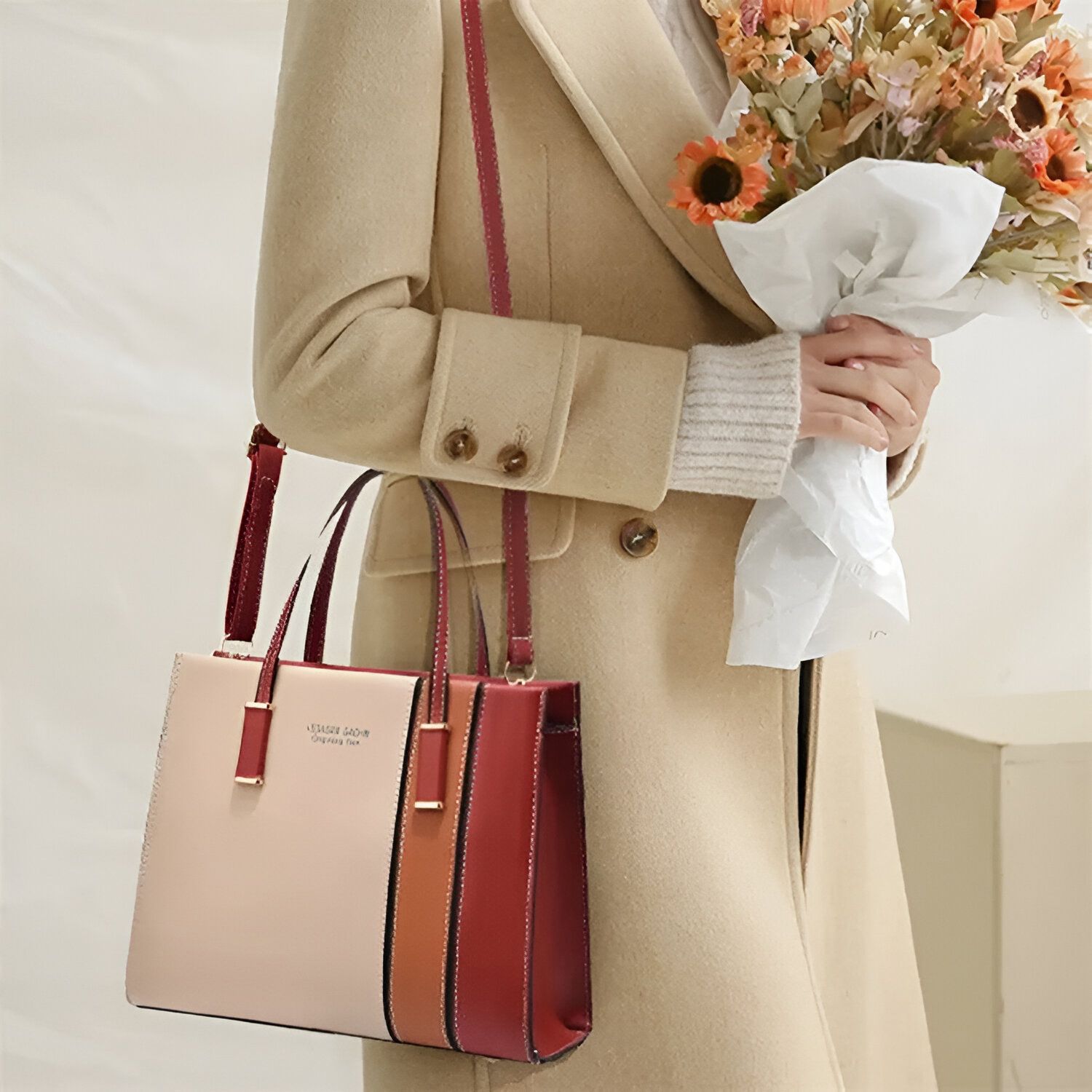 Une femme portant un manteau beige et tenant un bouquet de fleurs porte un sac à main en cuir rouge, beige et orange.