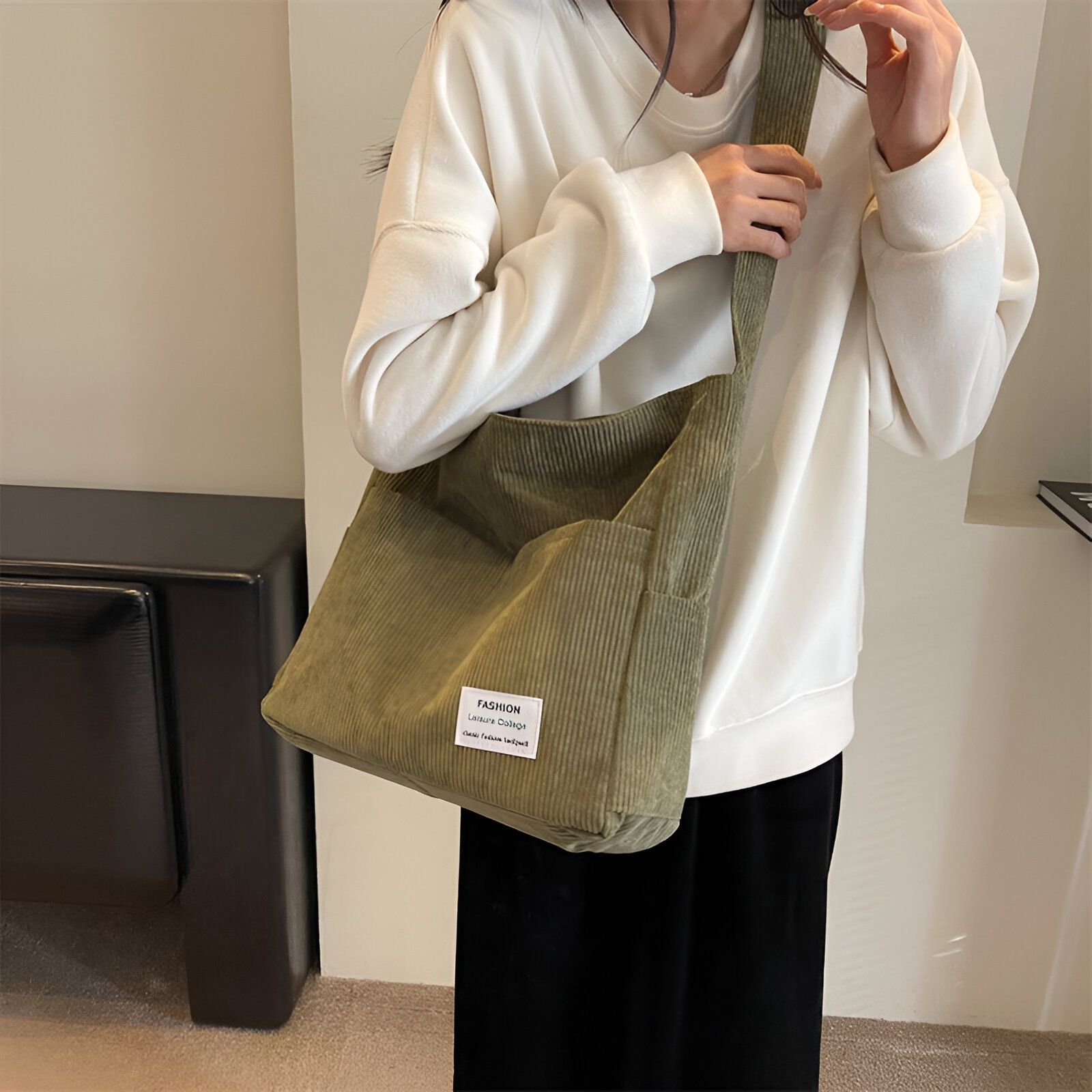 Une femme portant un sac à main en velours côtelé vert.  Le sac a une poche sur le devant et un étiquette qui dit "Fashion Leisure College".  La femme porte un chandail blanc.