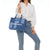 Une femme porte un sac à main en jean bleu avec l'inscription "THE TOTE BAG" en blanc. Elle porte un chemisier blanc et un jean bleu.