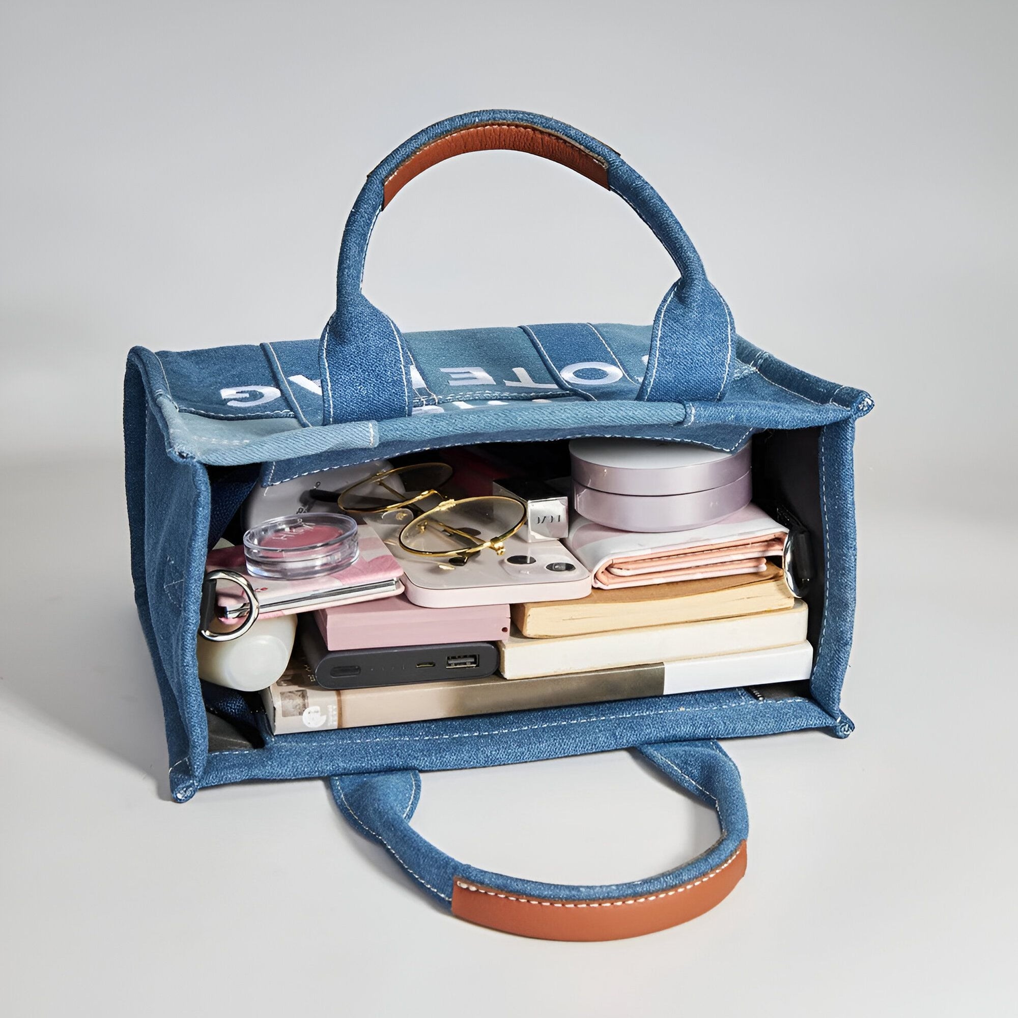 Un sac à main en jean bleu avec des anses en cuir marron, ouvert, montrant son contenu : lunettes, téléphone, maquillage et livres.