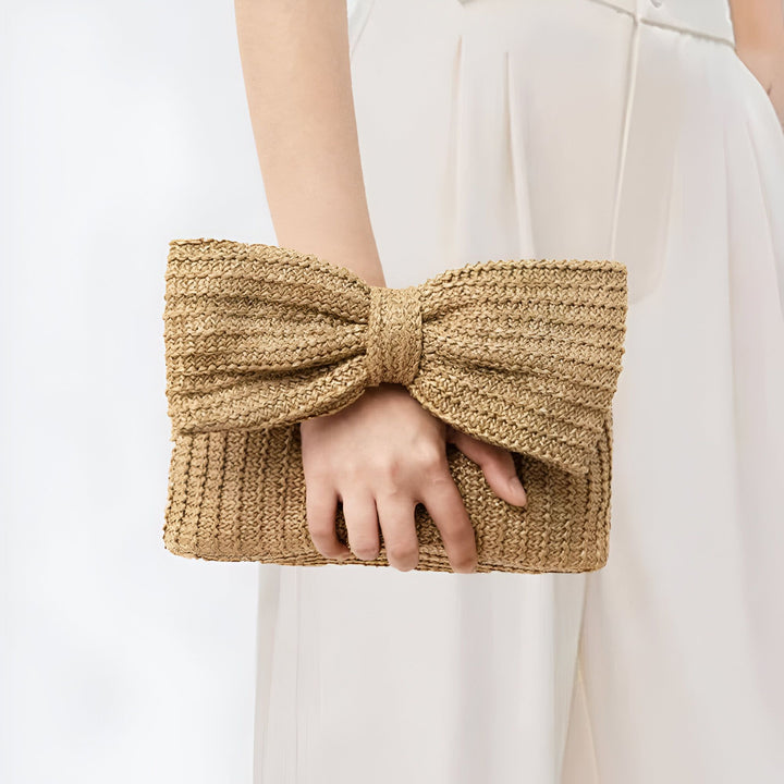Main tenant une pochette en paille marron clair avec un nœud sur fond blanc., 46194021794016