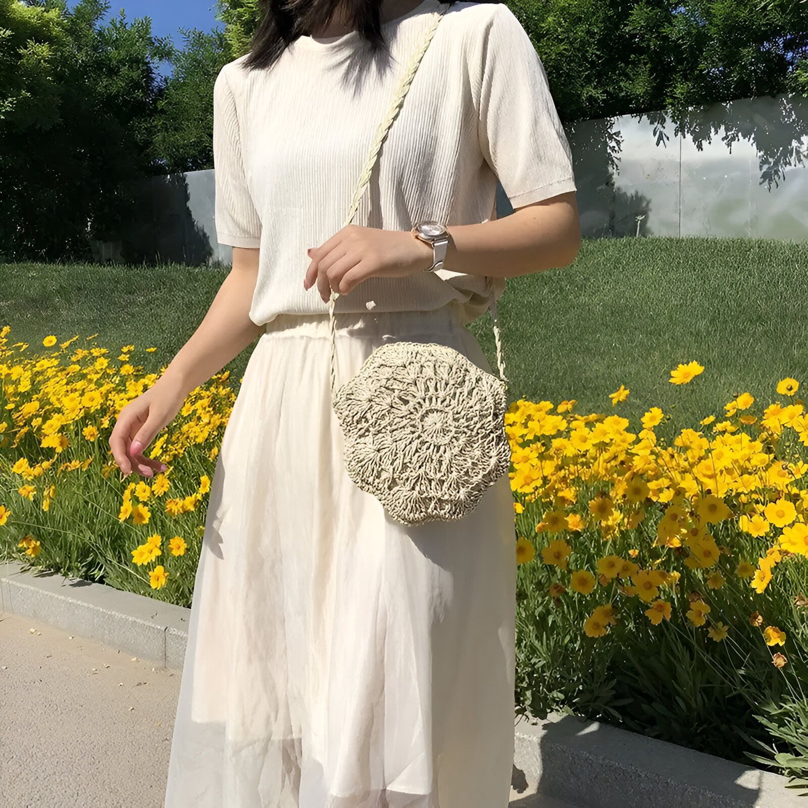 Une femme portant une robe blanche et un sac à main rond en paille, debout devant un lit de fleurs jaunes.