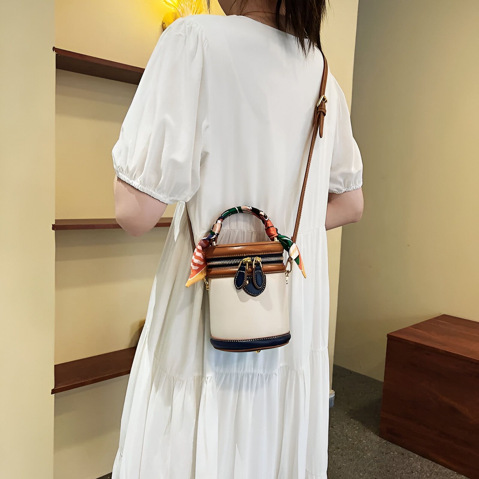 Une femme portant une robe blanche et un petit sac à main cylindrique avec une bandoulière en cuir marron. Le sac a une fermeture éclair en métal doré et une garniture en cuir bleu foncé. Le sac est décoré d'un foulard coloré.