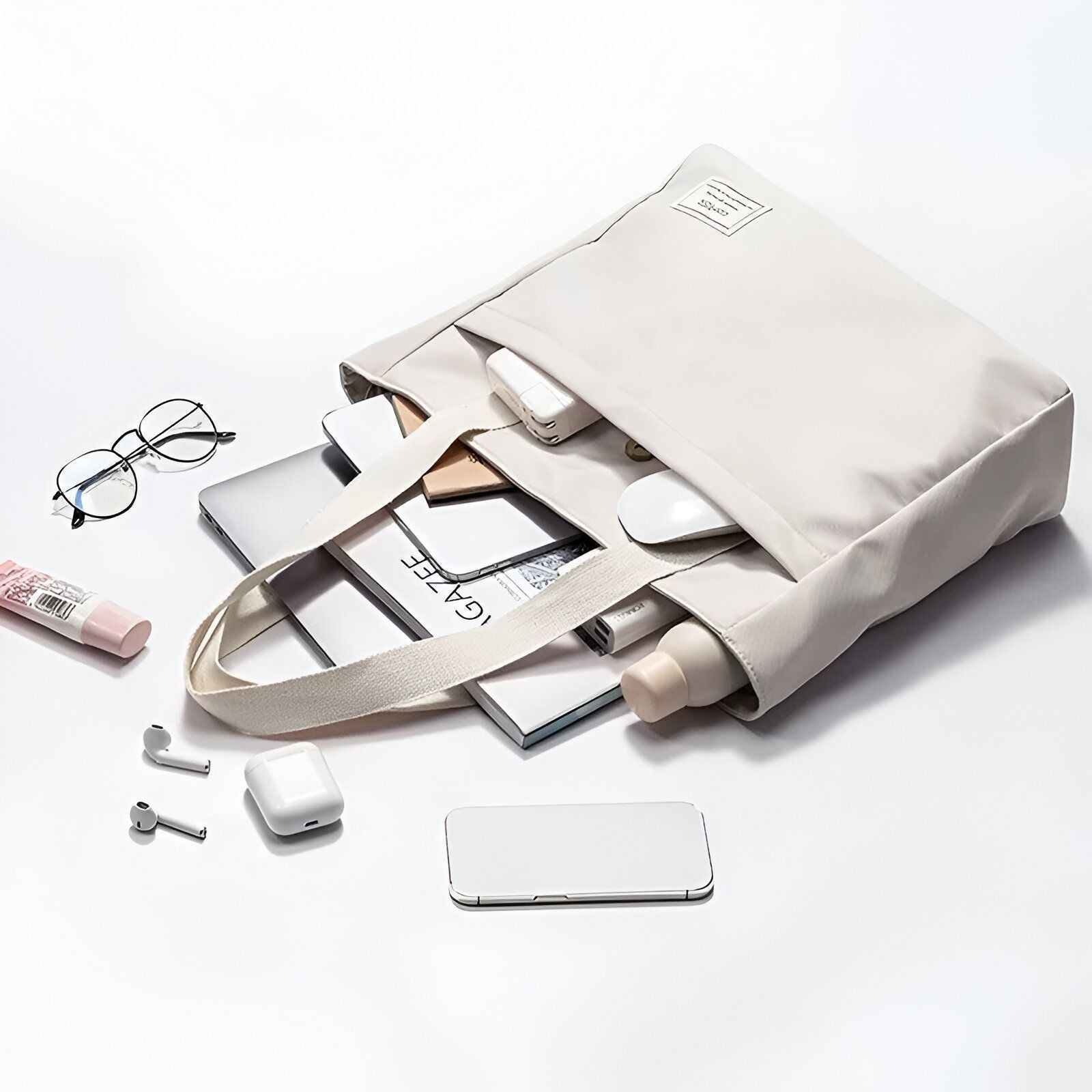 Un sac fourre-tout beige ouvert avec un ordinateur portable, un magazine, une bouteille, des écouteurs et une souris à l'intérieur.  Il y a aussi des lunettes et une bouteille de lotion sur le côté.