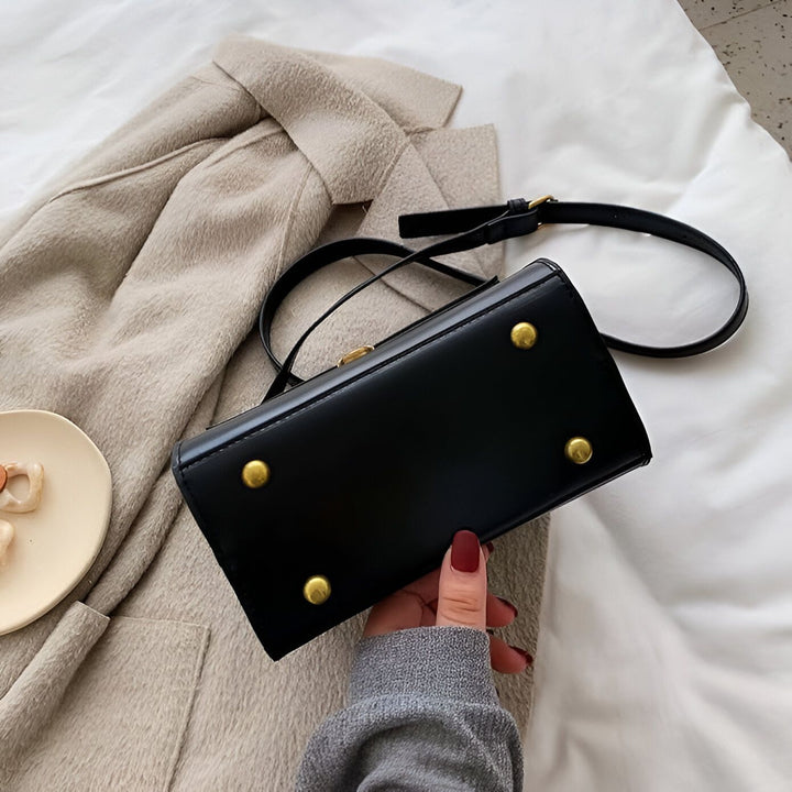 Sac à main noir à base cloutée dorée, tenu par une main, près d'un manteau beige., 46194026971360,46194027004128,46194027036896