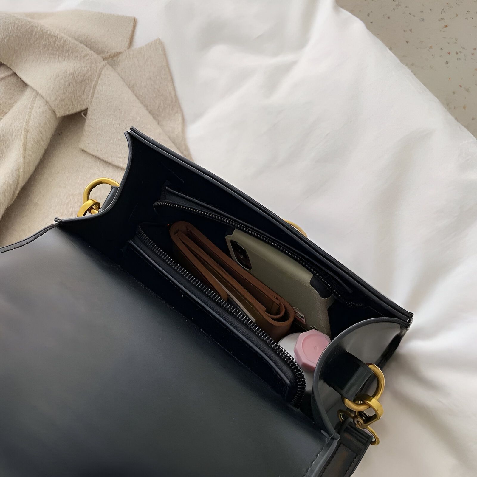 Un sac à main noir ouvert avec un téléphone, un portefeuille et un petit flacon à l'intérieur.  La fermeture éclair est ouverte.