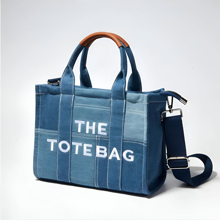 Sac fourre-tout en jean bleu avec anses en cuir marron et bandoulière. Texte blanc "THE TOTE BAG"., 46193991057632