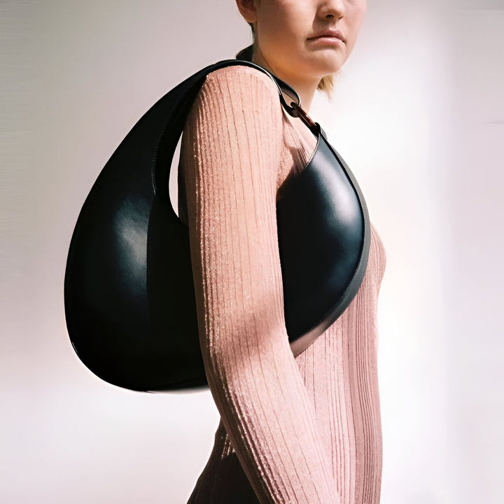 Sac noir asymétrique en cuir porté épaule par une femme en tricot rose., 46194017698016,46194017730784,46194017763552