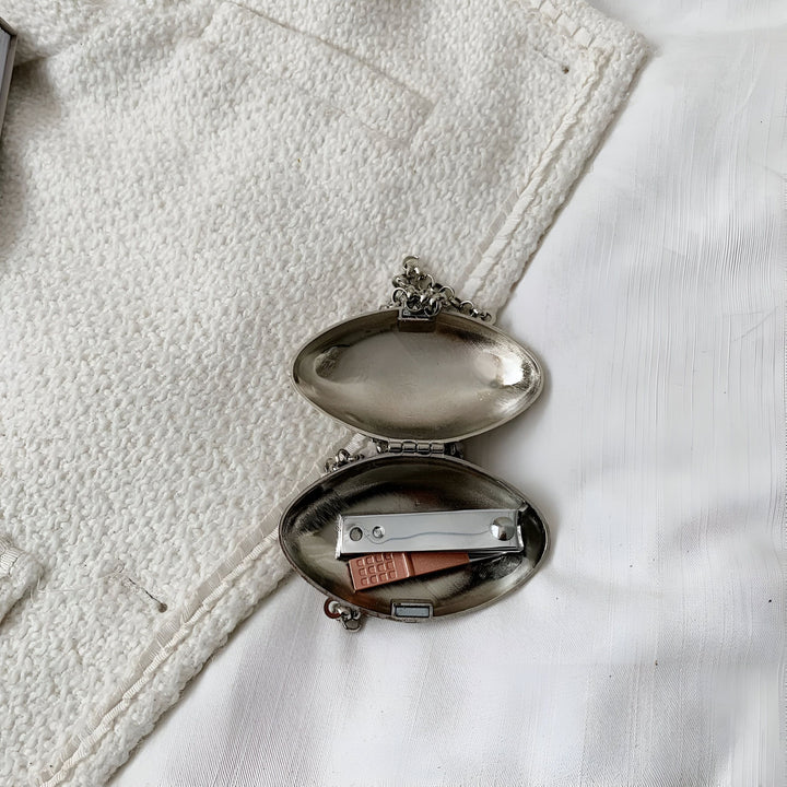 Boîte ovale métallique ouverte avec coupe-ongles et petit outil sur un tissu blanc texturé., 46193973788896,46193973821664
