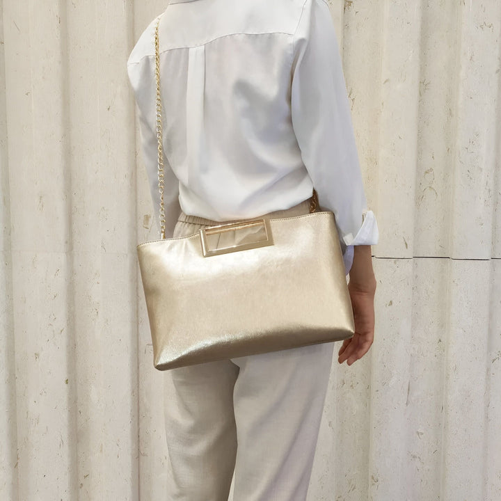 Vue de dos d'une personne portant une pochette dorée sur l'épaule. Chemise blanche, pantalon beige., 46194031853792